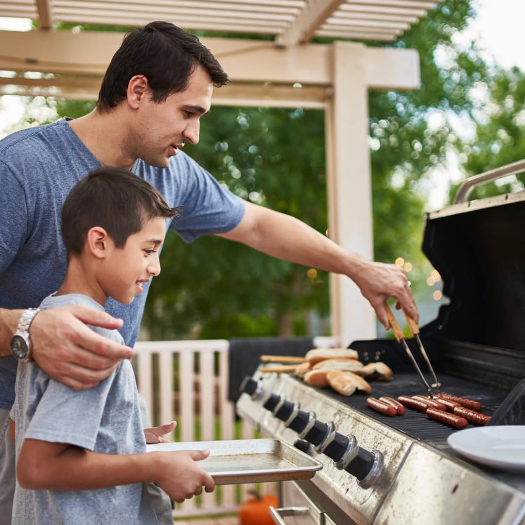 Adult grilling hot dogs and buns with a child who is holding a tray next to the grill.