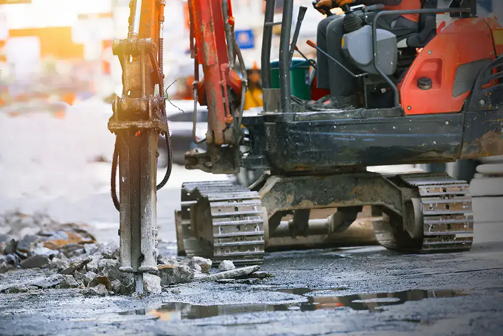 Excavator with hydraulic breaker breaks up asphalt on a city street.