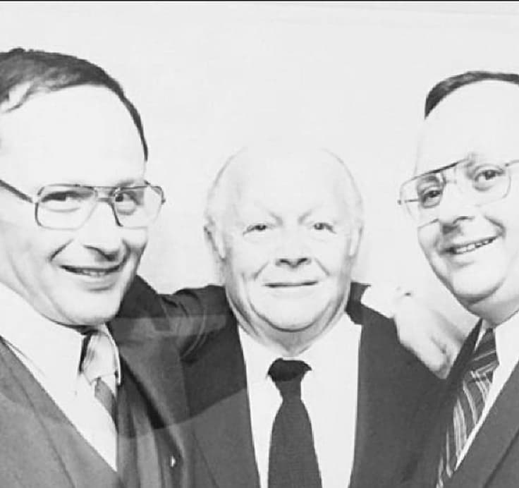 Three men in suits smile together in a black and white photo, standing closely in a formal setting.
