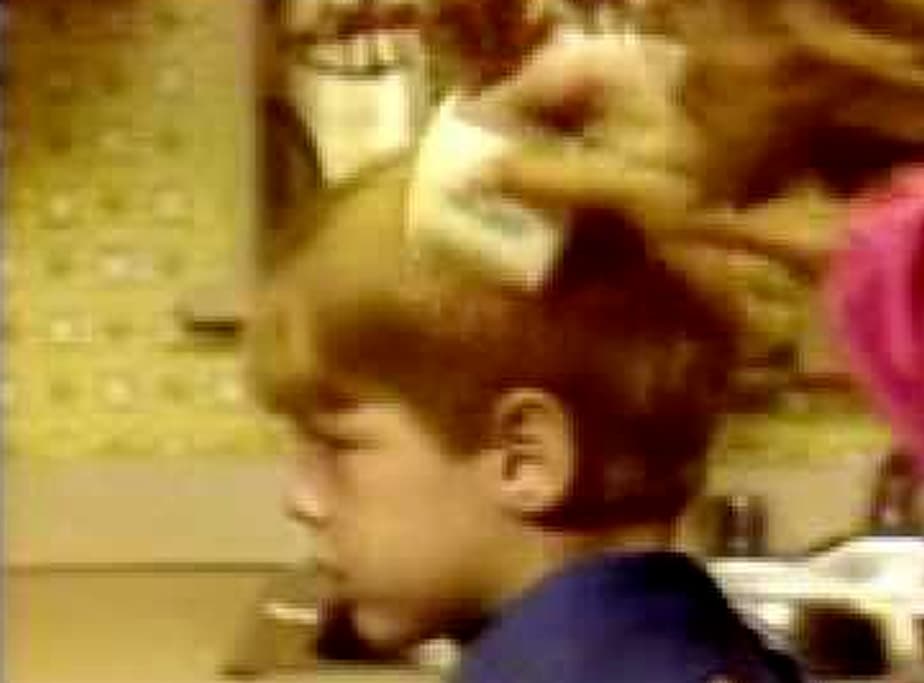 Boy sits as someone brushes his hair at home; patterned wall visible in the background.